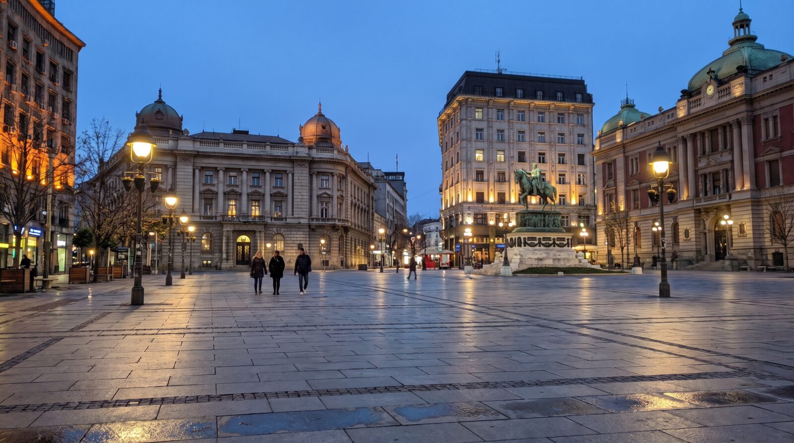 A Belgrade city square in the evening, lit by streetlamps with a few pedestrians crossing